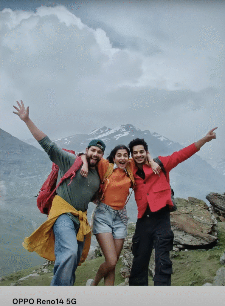 A joyful trio of young trekkers—two men and one woman—stand arm-in-arm on a grassy mountain ledge, backpacks on, raising their free arms against a backdrop of cloud-shrouded, snow-capped peaks.