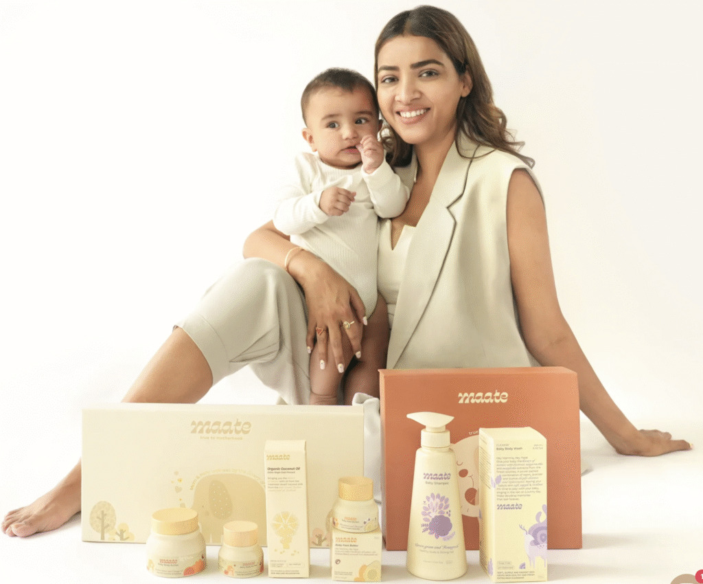 Priyanka Raina in a cream waistcoat holding her infant son beside an array of Maate baby-care products displayed against a white backdrop