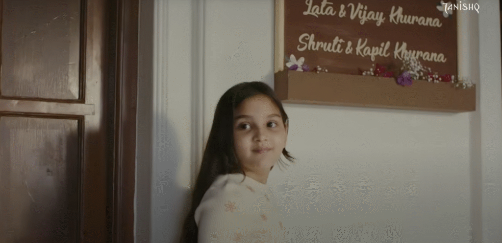 A girl  stands in front of a white wall inside a home, with a wooden nameplate above her that reads “Lata & Vijay Khurana, Shruti & Kapil Khurana,” capturing a moment from Tanishq’s Raksha Bandhan campaign about inclusive family spaces.