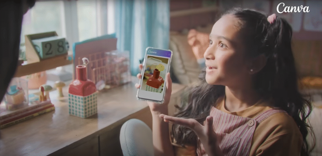 Young girl using Canva mobile app on smartphone displaying colorful digital design while sitting at wooden table with craft supplies and gift boxes in background, demonstrating intuitive mobile design interface from Dil Se Design Tak campaign