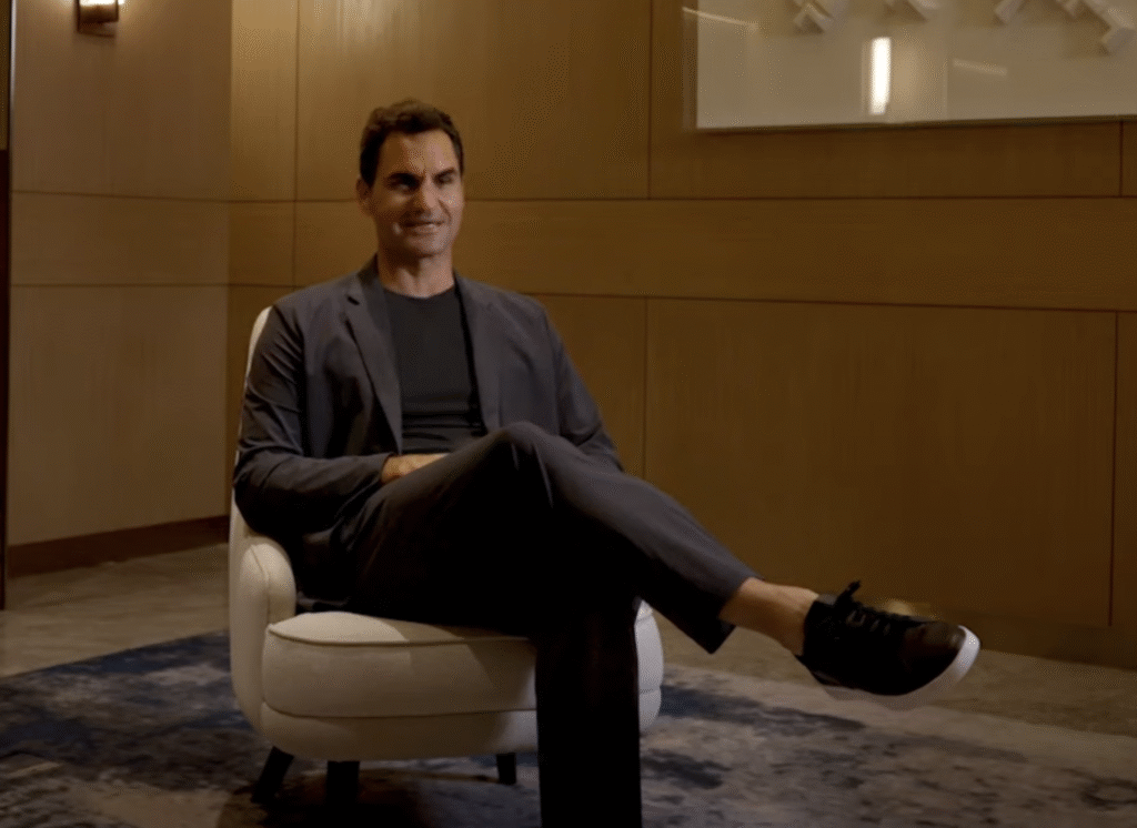 Federer in a Minimalist Setting 
Roger Fedrer in a dark suit sits cross-legged in a modern, softly lit room, exuding poise and understated luxury.