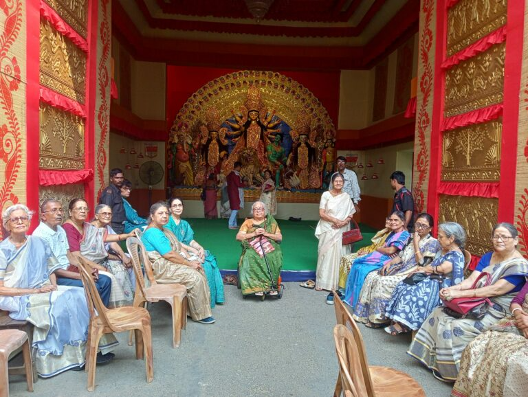 Senior citizens gathered in traditional attire sitting in chairs around an ornate Durga Puja pandal, representing diverse age demographics often overlooked in tech marketing campaigns