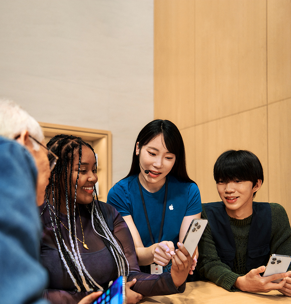 Apple Store specialist demonstrating iPhone features to diverse customers around a wooden table, engaging an elderly man, a young woman, and a teenager in hands-on product exploration.